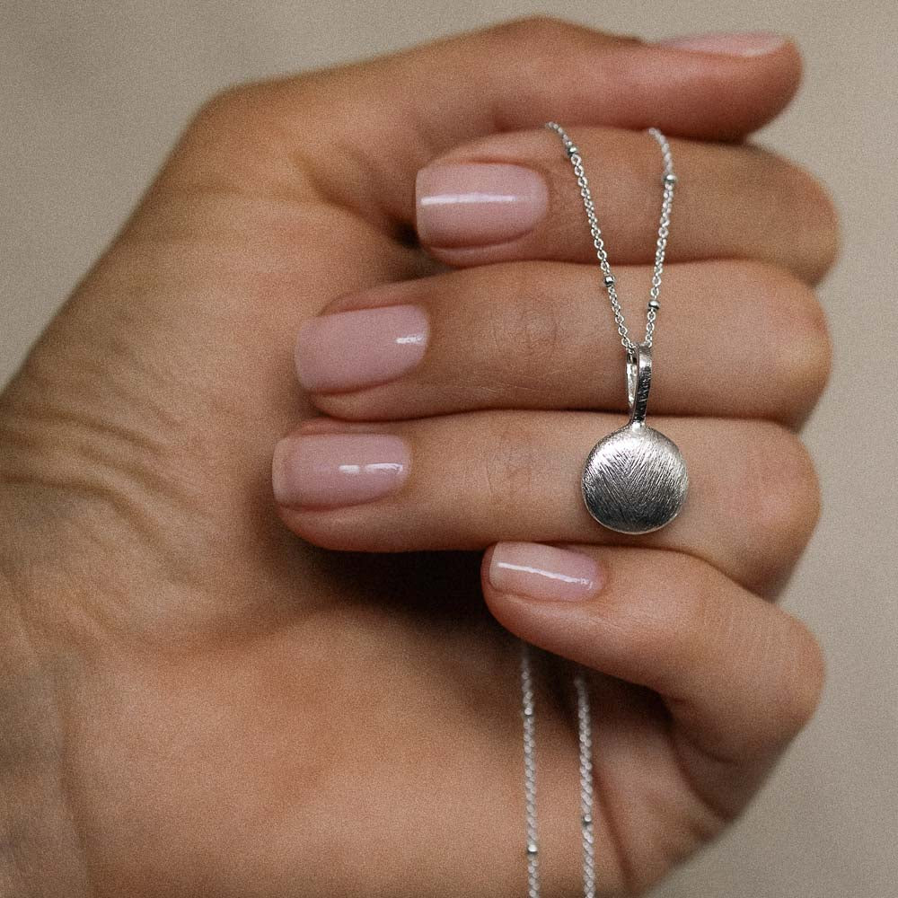 a brushed silver disc pendant hanging from a silver necklace chain held in a womans hands from Inesce Jewellery