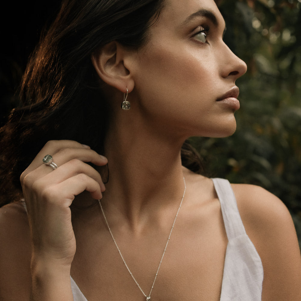 a woman wearing a pair of green amethyst square faceted earrings  and ring in lush greenery