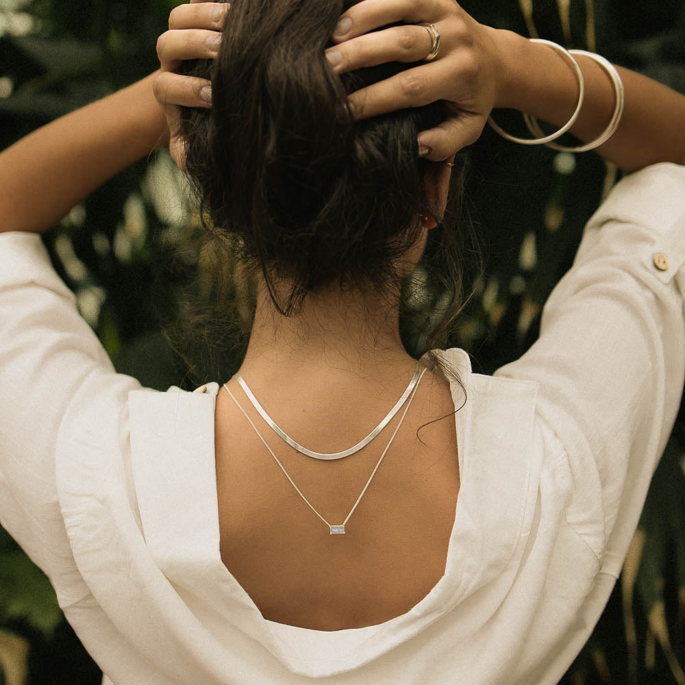 an earthy woman with her back facing us wearing silver bangles and a moonstone baguette necklace in a jungle with a white shirt on