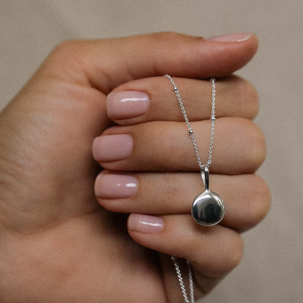 a polished silver disc pendant hanging from a silver necklace chain held in a womans hand by Inesce Jewellery