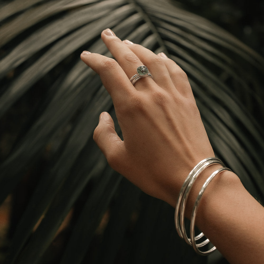a hand wearing a green amethyst silver ring and three silver bangles in a lush forest