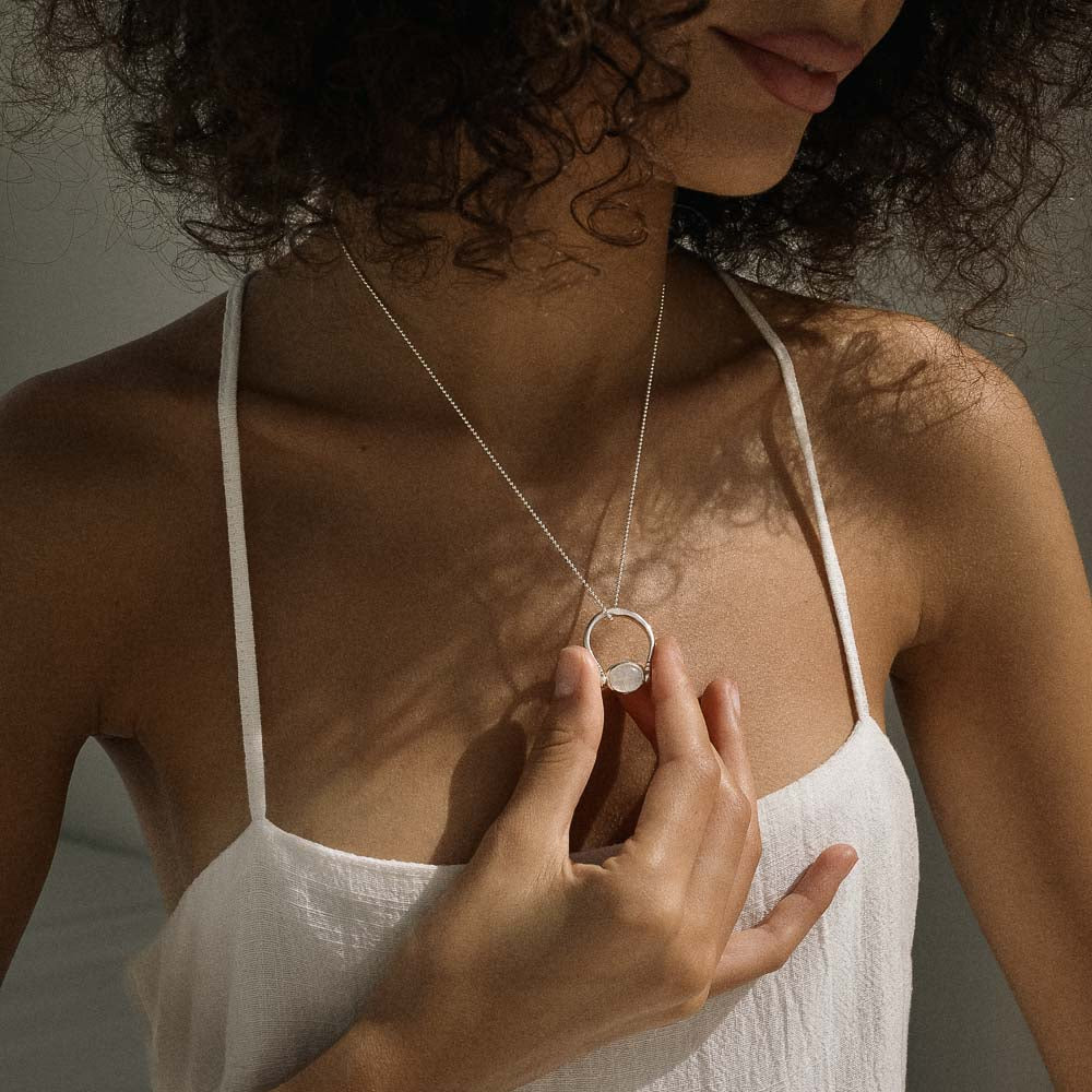 Woman wearing a white top and holding a necklace with a circular gem pendant.